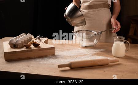 Woman sifting flour over a glass bowl, surrounded by baking ingredients like eggs, milk, and a rolling pin Stock Photo