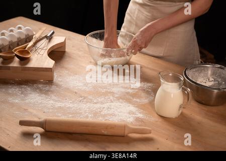Person kneading dough for baking homemade bread on a rustic wooden table with flour, milk, and eggs Stock Photo