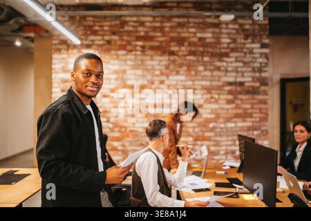 Man stands in office with papers while colleagues work at desks in a modern workplace environment Stock Photo