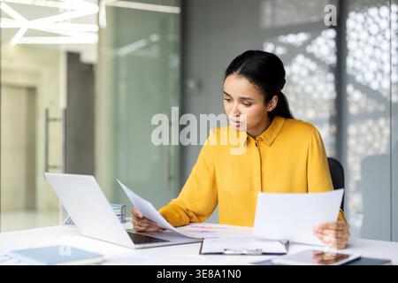 Young professional businesswoman analyzing important financial reports and paperwork while sitting at a desk with a laptop in a contemporary office environment Stock Photo