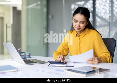 Young professional woman or accountant sitting at an office desk, analyzing financial documents and using a calculator while working with a laptop and other paperwork, managing business finances Stock Photo