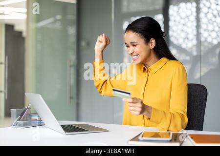 Young woman celebrates successful online purchase, holding a credit card and raising her fist while working at a laptop on a tidy office desk, excited and satisfied with the transaction Stock Photo