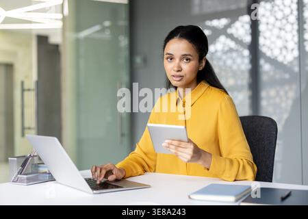 Young professional woman sitting at her desk, working efficiently with a laptop and holding a digital tablet in a modern office environment, demonstrating productivity and business connectivity Stock Photo