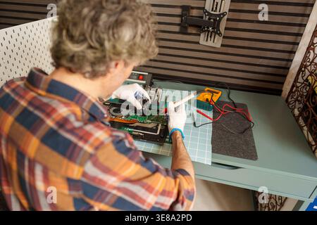 Man wearing gloves working on an open laptop motherboard, testing circuits with a multimeter on a workbench Stock Photo