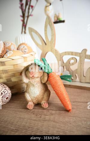 Easter bunny figurine holding a carrot next to a basket of decorated eggs on a wooden table. Stock Photo