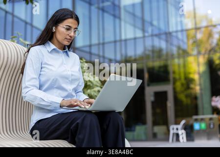 Young businesswoman typing on a laptop computer while sitting on a modern bench outside a glass office building, embracing flexible work and global connectivity Stock Photo