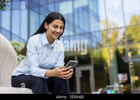 Young businesswoman smiling, sitting outdoors and using a mobile phone, representing remote work, digital communication, and modern technology in a corporate environment Stock Photo