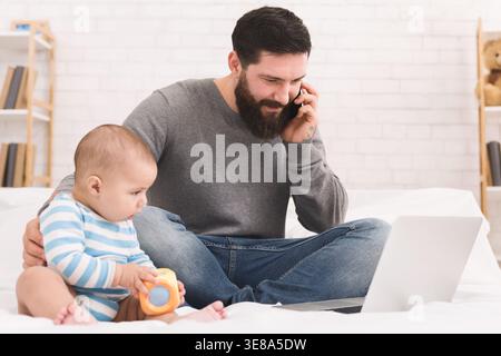 Father Talks on Phone While Sitting With Baby on Bed and Using Laptop in Home Setting Stock Photo