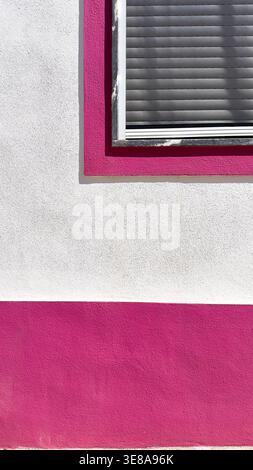 abstract Architectural detail of a white stucco building with vibrant pink painted accents and a window with blinds in vertical composition portugal Stock Photo