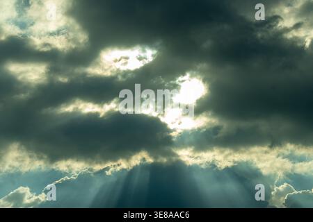 A dramatic, moody sky with dark stormy clouds dramatically parting to reveal powerful sun rays bursting through, casting ethereal light beams across t Stock Photo