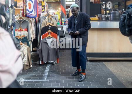 London, UK, 5  December 2026,  Senior man in a stylish dark coat and beanie checking his phone while walking past a vintage clothing stall at Spitalfi Stock Photo