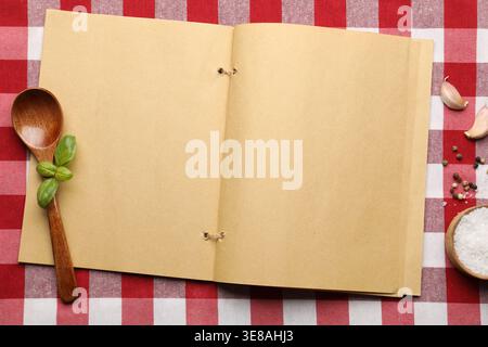 Recipe book, different spices and wooden spoon on checkered tablecloth, flat lay Stock Photo