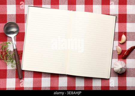 Recipe book, different spices and spoon on checkered tablecloth, flat lay Stock Photo