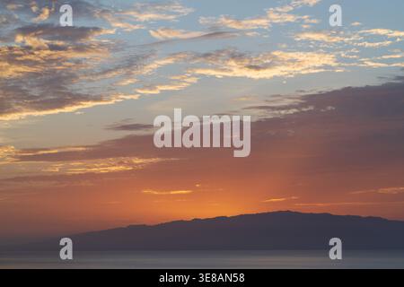The sky is painted with hues of orange, pink, and blue as the sun dips below the horizon, casting a silhouette of distant mountains over the calm sea Stock Photo