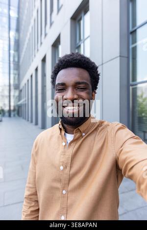 Confident young man with curly hair and beard smiling at camera outside a modern office building, casual entrepreneur portrait conveying success, friendliness, and urban optimism Stock Photo