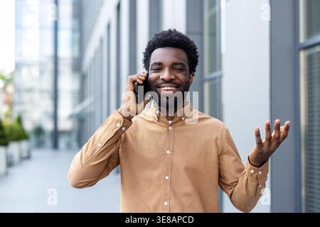 Smiling young professional walking through a modern city, talking and gesturing on his smartphone, confident and relaxed while handling business communication outdoors Stock Photo