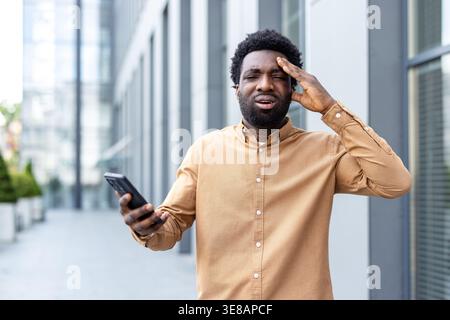 Young adult man experiencing headache and frustration while holding a phone outdoors, representing stress, financial problems, and business challenges Stock Photo