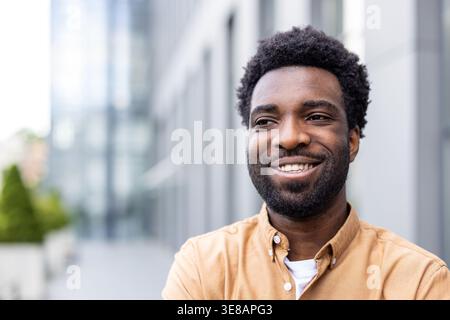 Young man with a warm smile looking away, expressing happiness and confidence while standing outdoors in a modern city environment, conveying professionalism and success Stock Photo
