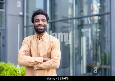 African man standing outdoors with arms crossed, projecting confidence, success, and professionalism against a modern glass building, representing entrepreneurship and business achievement Stock Photo