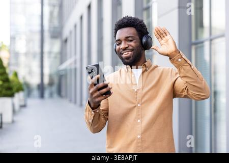 Young man smiling and waving during a video call on his smartphone, wearing headphones and standing on an urban street, enjoying modern communication outdoors Stock Photo