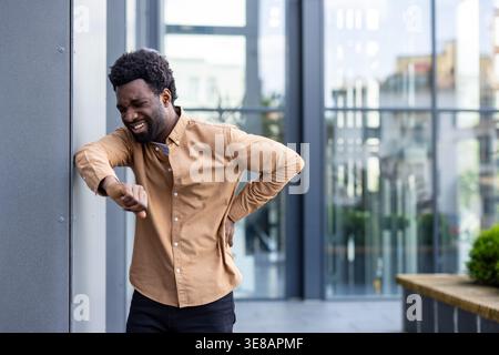 Young adult man in brown shirt clutching lower back and grimacing against a building wall, showing visible pain and discomfort from a spine or lower back strain in an urban setting Stock Photo