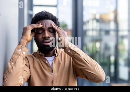 Young man experiencing severe headache and migraine, pressing hands to temples, feeling exhausted and overwhelmed with mental pressure, suffering from stress and burnout while standing outdoors Stock Photo