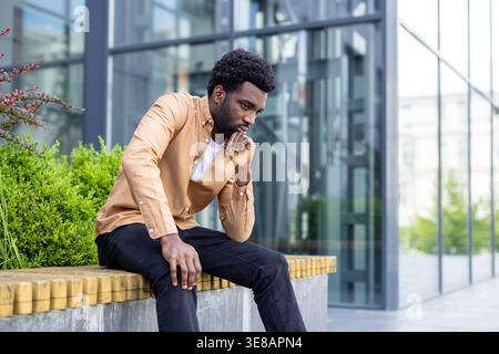 Thoughtful young man sitting outdoors on a modern city bench, resting his chin on his hand while contemplating and reflecting on challenging issues, looking pensive and concerned Stock Photo