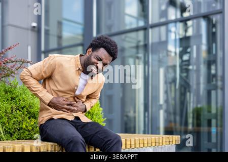 Young adult man experiencing severe abdominal cramps and discomfort while sitting on a bench in an urban environment, clutching his midsection with a pained expression Stock Photo