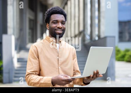 Man holding a laptop and smiling at camera, representing concepts of freelance work, online education, and modern entrepreneurship in an urban outdoor setting Stock Photo