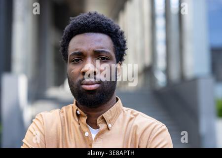 Young adult man with a serious expression and beard looking directly at the camera, portraying feelings of determination and confidence while standing in an urban environment Stock Photo