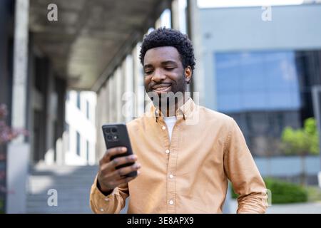 Young african descent man smiling and holding a mobile phone in his hands, engaging with digital content while standing in an urban environment during the day Stock Photo