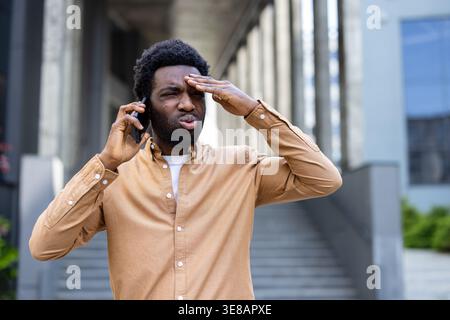 Young man feeling disappointed and frustrated, experiencing stress and headache while having a difficult phone conversation outside a modern city building, reacting to bad news Stock Photo