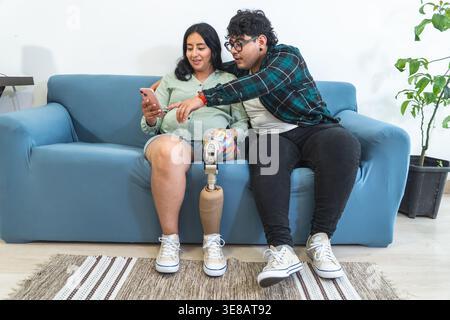 Pregnant woman with a prosthetic leg and partner using a smartphone together on a sofa at home Stock Photo