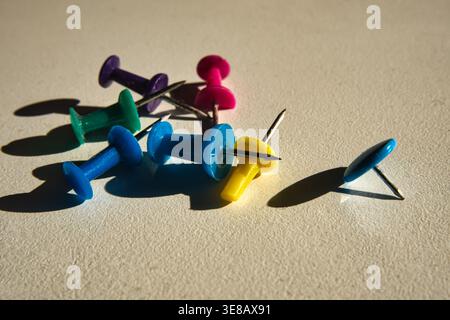 A group of vibrant multi-colored plastic pushpins scattered on a textured white surface, featuring dramatic long shadows under bright light. Business Stock Photo