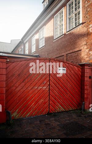 Weathered red wooden gate in front of classic brick building. A large, rustic red wooden gate with a diagonal plank pattern in front of a brick house. Stock Photo