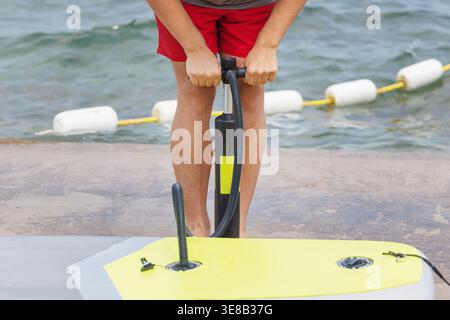 Fit young man stands on dock, pumping air into paddleboard, ready to enjoy a day on the water and explore the ocean. Stock Photo