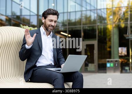 Smiling businessman wearing earbuds and suit, waving hand to laptop screen during an online video call for remote work or virtual meeting outside modern office building Stock Photo