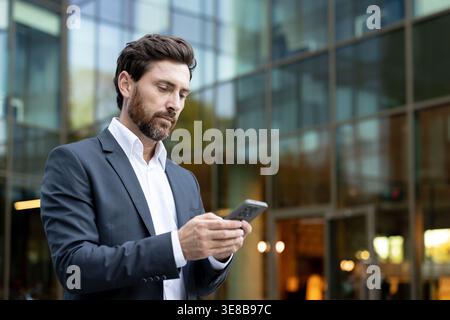 Businessman using smartphone for networking and corporate communication, standing outside a modern glass office building, representing mobile technology, connectivity, and urban professionalism Stock Photo