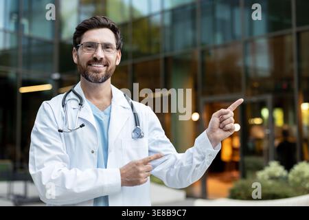 Smiling male doctor wearing a white lab coat, stethoscope, and glasses, confidently pointing for a medical presentation or advertisement in front of a modern hospital building Stock Photo