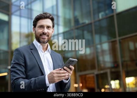 Professional man happily using a smartphone while standing outside a modern office building. Representing mobile communication. Business connectivity. And digital networking in a busy city environment Stock Photo