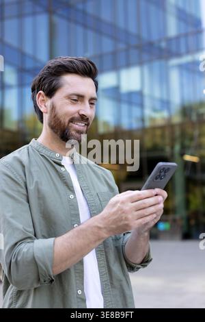 Happy man smiling while holding and interacting with a mobile phone, standing outdoors in a city setting with modern glass buildings in the background Stock Photo