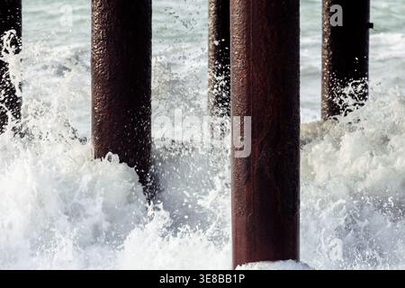 Strong sea waves hitting pier columns with splashing foam and water droplets. Close-up coastal scene capturing motion, energy and interaction between Stock Photo