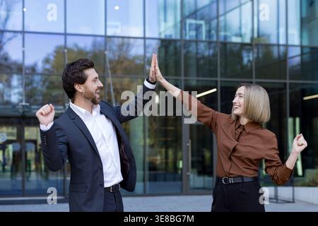 Business colleagues celebrating a successful project achievement, giving a high five with joyful expressions in front of a modern office building, symbolizing teamwork and partnership Stock Photo