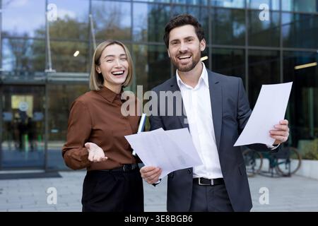 Two cheerful business professionals, man and woman, standing outside a modern corporate building, holding documents and smiling, conveying teamwork, success and confident collaboration Stock Photo
