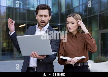 Two stressed business partners looking at a laptop with confused, frustrated expressions while encountering a technical problem or system error outside a modern office building Stock Photo