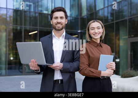 Diverse business partners stand confidently before a modern glass office tower, holding a laptop and tablet to illustrate teamwork, innovation and professional collaboration in urban corporate life Stock Photo