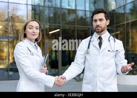 Healthcare professionals, a male and female doctor, shaking hands in front of a modern hospital building, symbolizing agreement, cooperation, and successful teamwork in medicine Stock Photo
