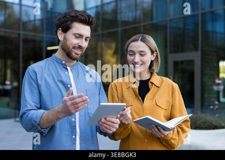 Business professionals engaging in an informal outdoor meeting, discussing work and ideas using a digital tablet and notebook in front of a modern office building Stock Photo