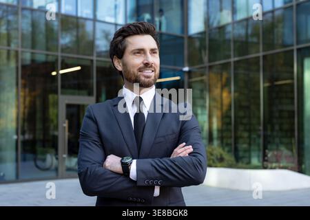 Confident businessman in a dark suit with crossed arms standing outdoors in front of a modern glass office building, smiling and looking away, representing success and professionalism Stock Photo