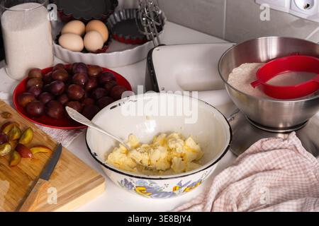 Cook is preparing to make plum pie in a home kitchen. Ingredients like plums, flour, sugar, and butter are on the counter. Tools for mixing and baking are nearby. Stock Photo